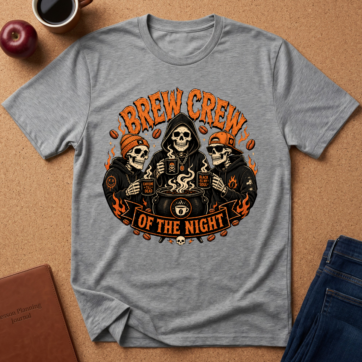 A heather gray short-sleeve graphic T-shirt laid flat on a cork surface, featuring three illustrated skeleton characters in black hoodies holding coffee mugs under orange text that reads 'BREW CREW OF THE NIGHT.' Surrounding props include a cup of coffee, an apple, a notebook, and folded blue jeans.