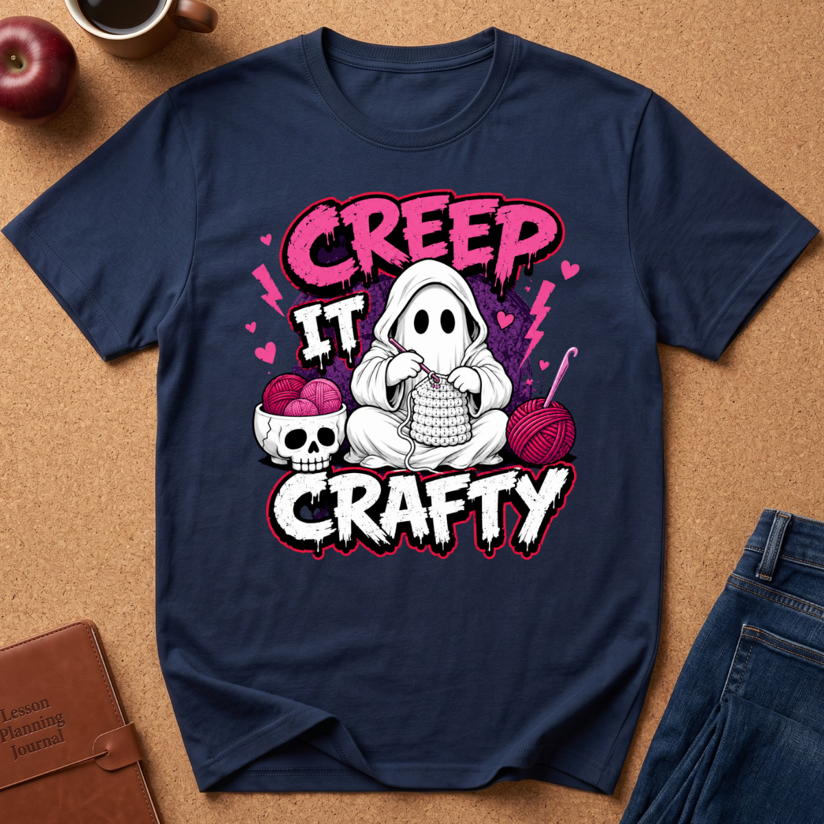 A navy blue graphic t-shirt laid flat on a brown cork background, featuring a cute cartoon ghost knitting with yarn, a skull bowl of pink yarn balls, and the text 'CREEP IT CRAFTY' in pink and white.