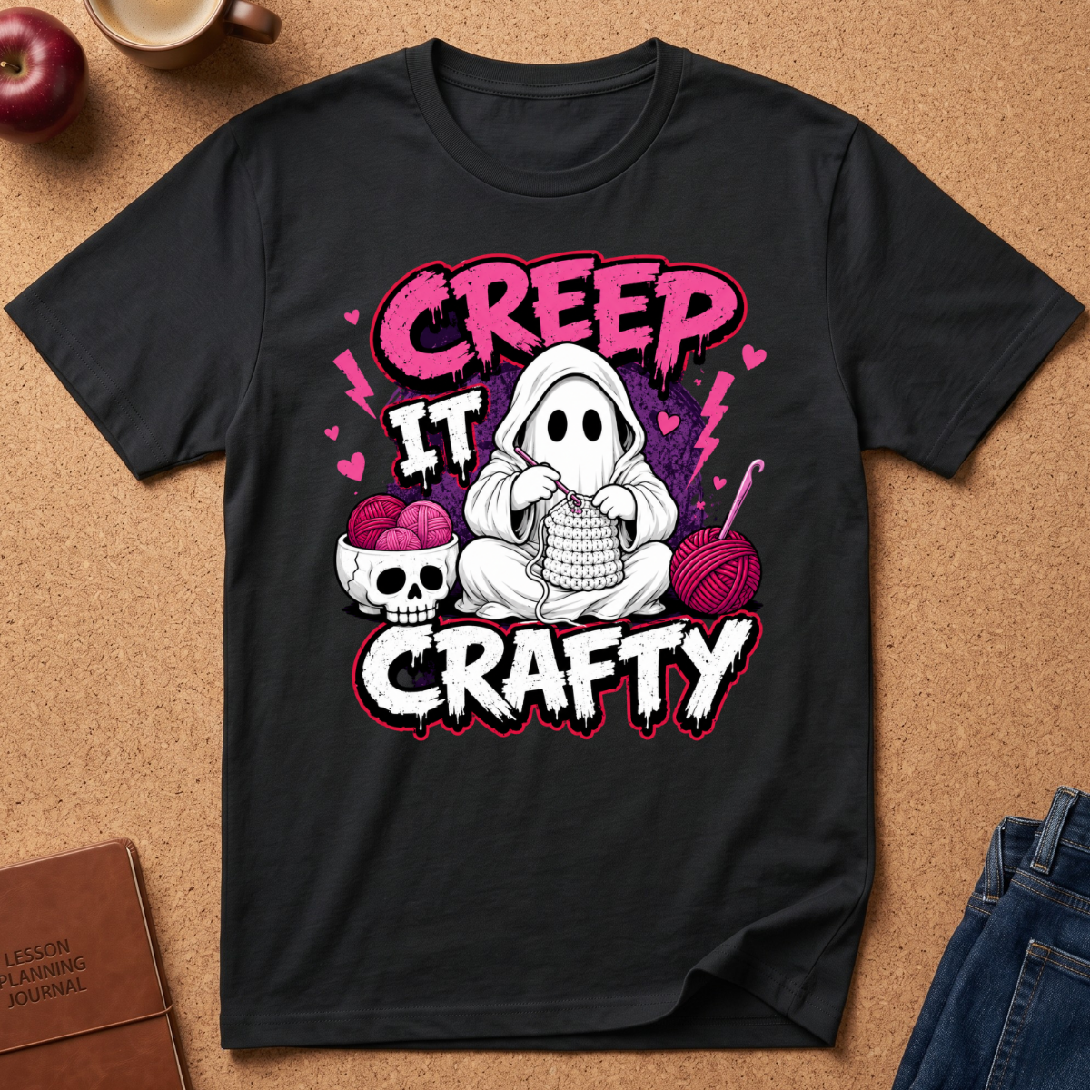 A black graphic t-shirt laid flat on a brown cork background, featuring a cartoon ghost knitting with pink yarn and the text 'CREEP IT CRAFTY' in pink and white. Styled flat-lay props include an apple, a coffee cup, a journal, and jeans.
