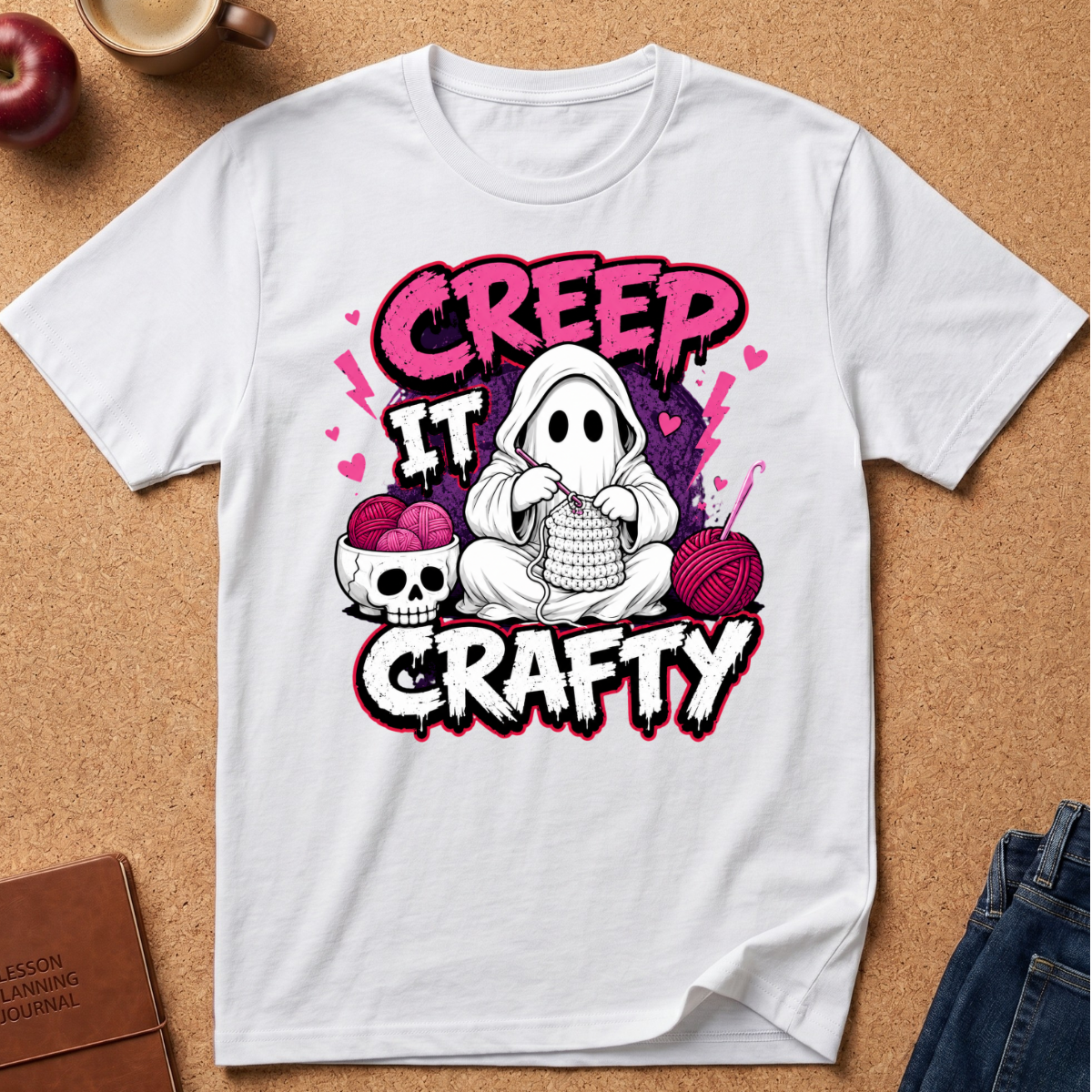 A white short-sleeve graphic T-shirt laid flat on a cork background, featuring a Halloween-themed design with the words 'CREEP IT GRAFTY' and an illustration of a ghost knitting, a skull with pink yarn, and pink hearts.