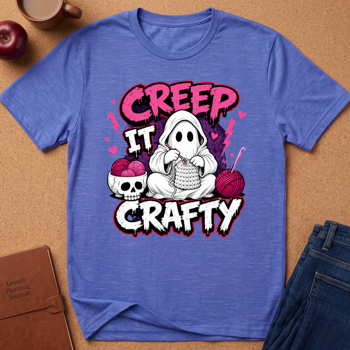 A heathered blue short-sleeve t-shirt laid flat on a cork background, featuring a Halloween-themed graphic with the words 'CREEP IT CRAFTY' and an illustration of a ghost knitting with yarn, a skull, and pink balls.