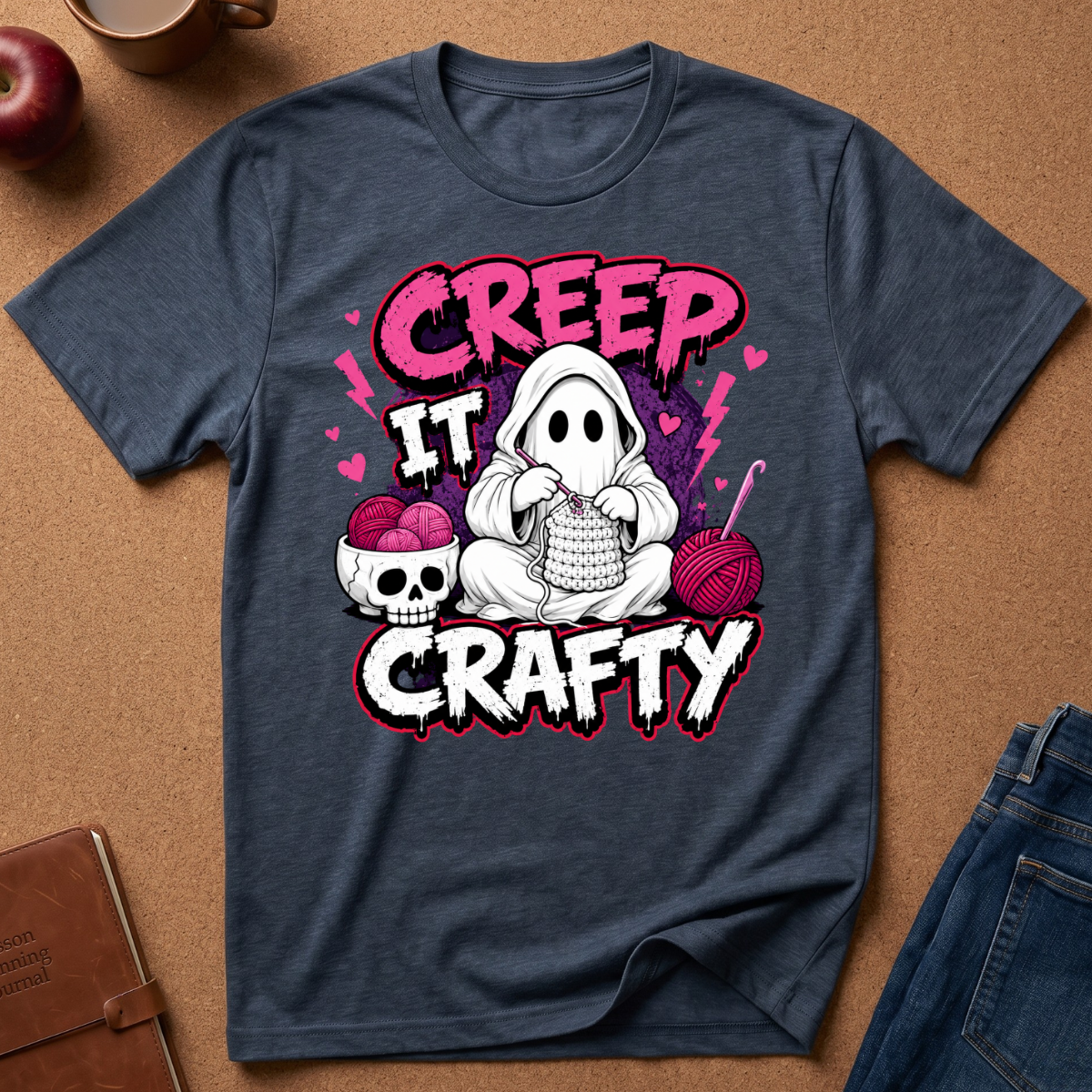 A navy blue graphic t-shirt laid flat on a brown cork background, featuring a cute cartoon ghost knitting with yarn, a skull, and the text 'CREEP IT CRAFTY' in pink and white.