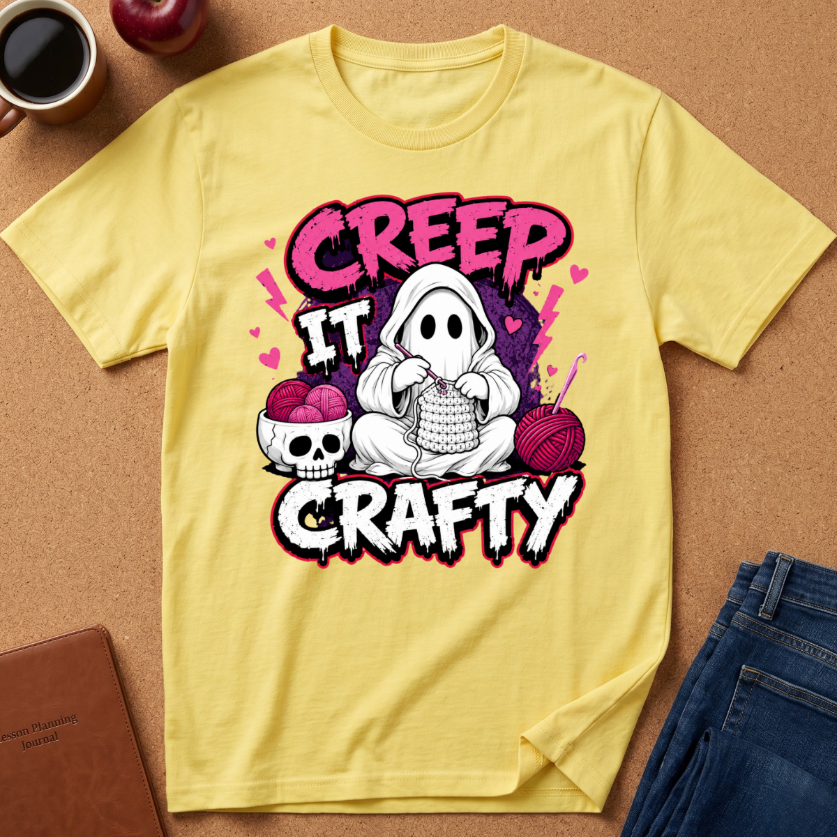 A yellow graphic t-shirt laid flat on a cork background with lifestyle props including a coffee mug, apple, notebook, and jeans. The t-shirt features a cartoon ghost knitting with the text 'CREEP IT IT CRAFTY' in pink and white.