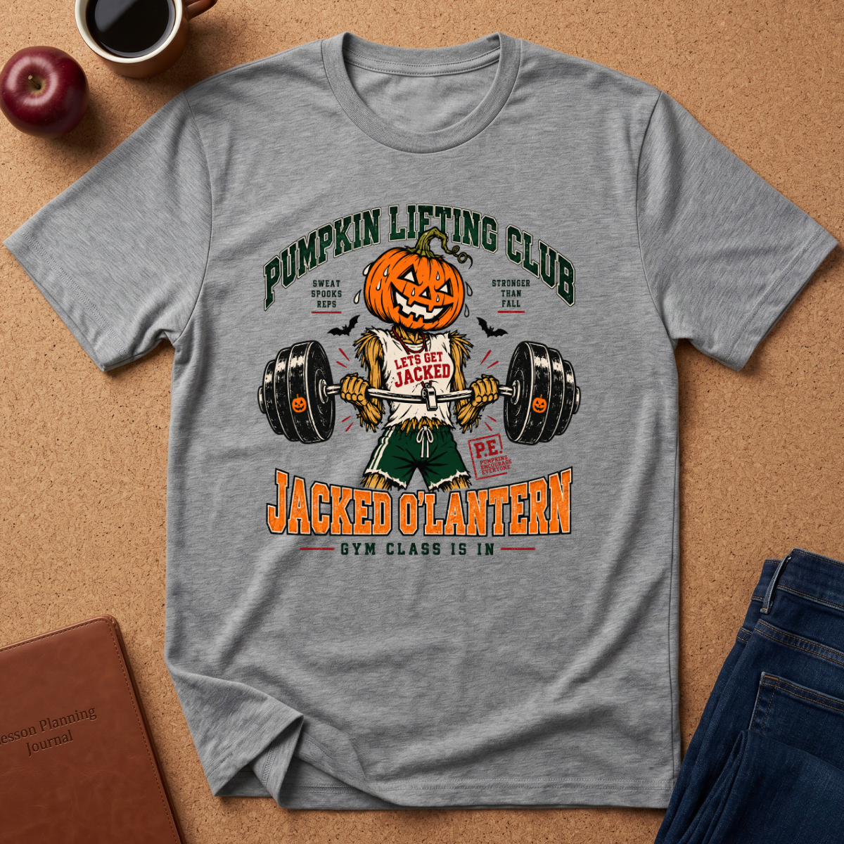 A heather gray graphic t-shirt laid flat on a cork background, featuring a Halloween-themed gym graphic with a jack-o'-lantern lifting a barbell and the text 'Pumpkin Lifting Club' and 'Jacked Olantern'.