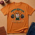An orange graphic t-shirt featuring a cartoon pumpkin lifting a barbell with the text 'Pumpkin Lifting Club' and 'Jacked Lantern'. The shirt is displayed in a flat-lay arrangement on a cork background with an apple, coffee mug, notebook, and jeans.