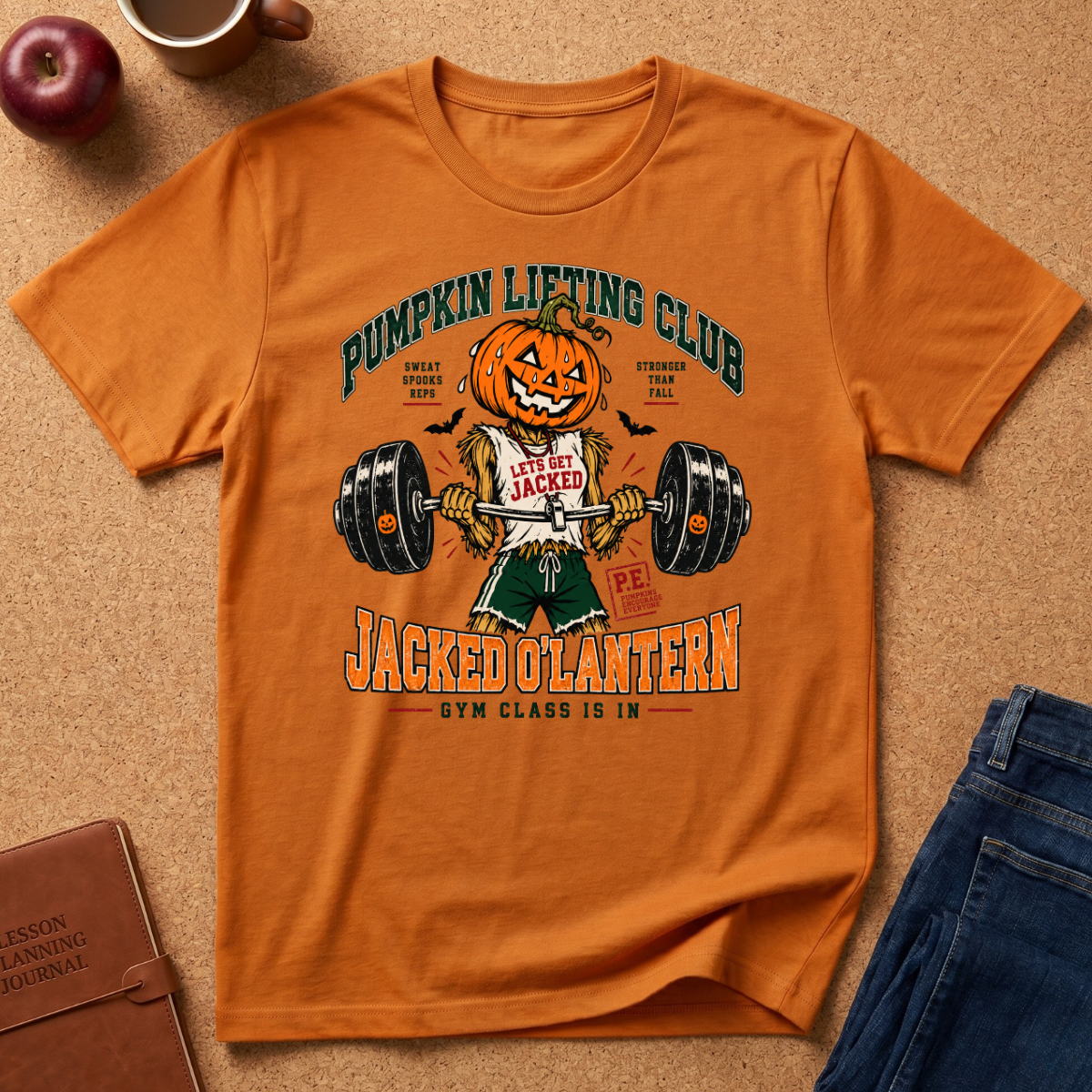 An orange graphic t-shirt featuring a cartoon pumpkin lifting a barbell with the text 'Pumpkin Lifting Club' and 'Jacked Lantern'. The shirt is displayed in a flat-lay arrangement on a cork background with an apple, coffee mug, notebook, and jeans.