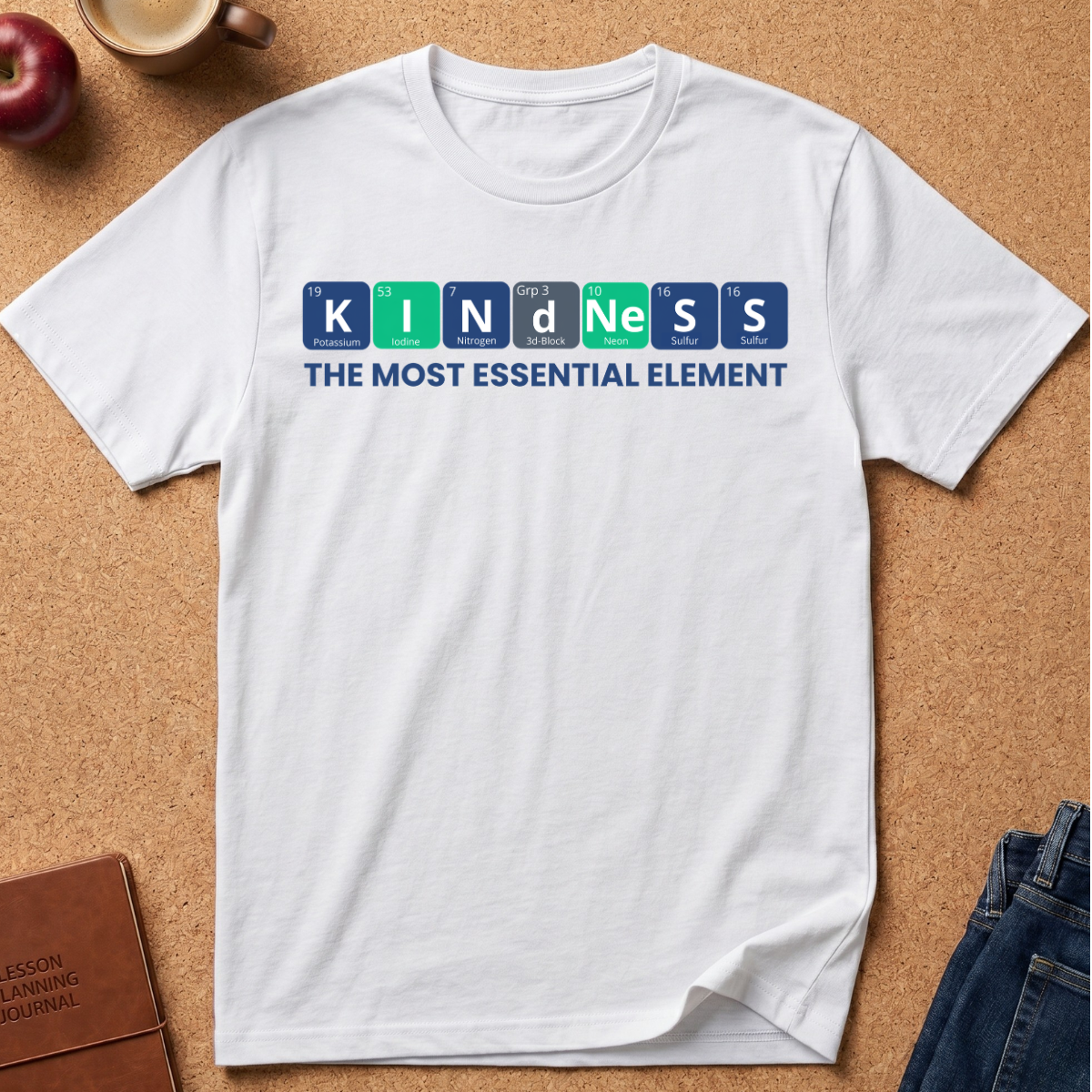 A white short-sleeve crewneck t-shirt laid flat on a cork background, featuring a graphic that spells 'KINDNESS' using Periodic Table-style letter tiles in blue, green, and gray, with the text 'THE MOST ESSENTIAL ELEMENT' underneath.