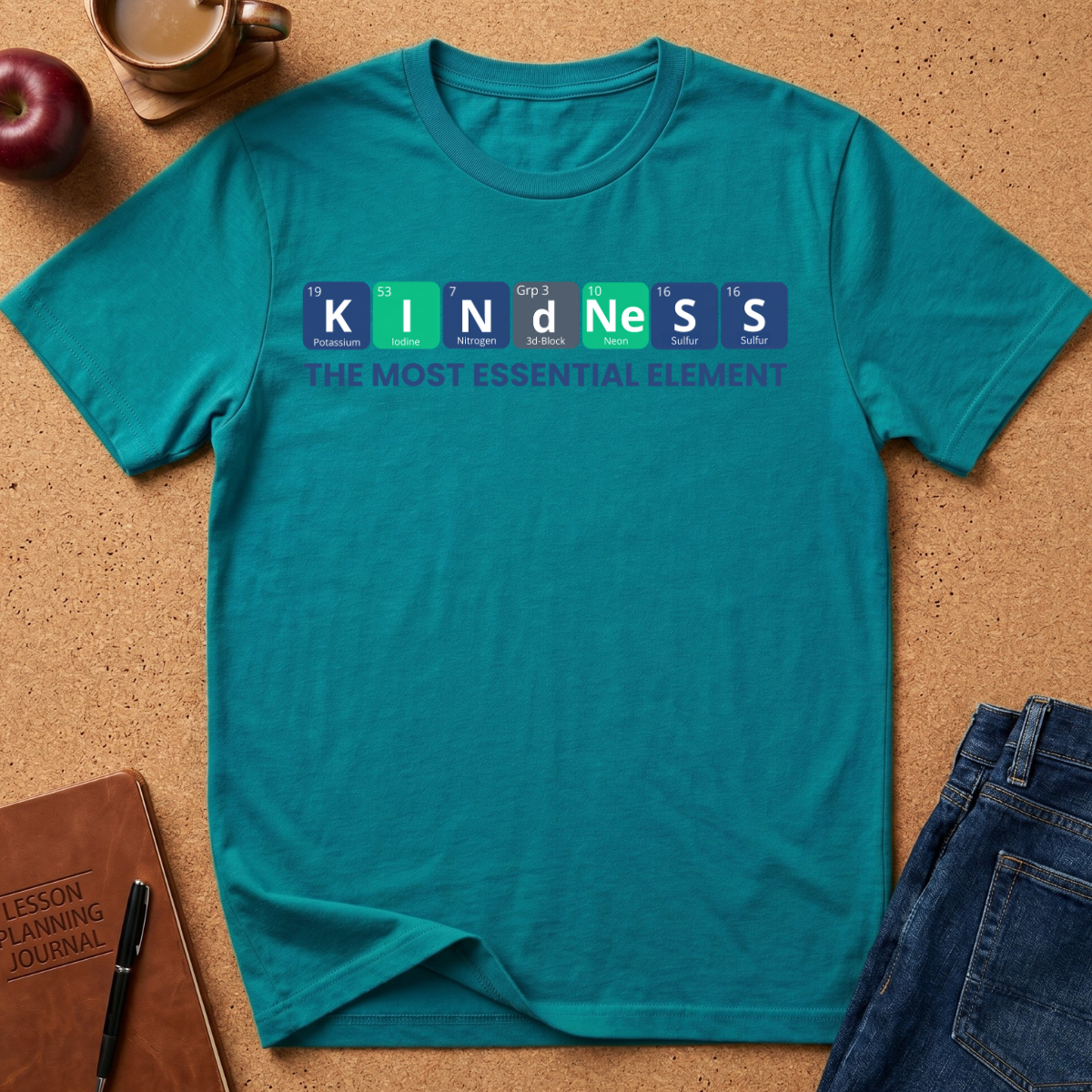 A teal short-sleeve t-shirt laid flat on a cork surface, featuring a periodic-table-style graphic that spells 'KINDNESS' with the phrase 'THE MOST ESSENTIAL ELEMENT' underneath. Styled flat-lay props include a brown notebook, pen, apple, coffee cup, and blue jeans.