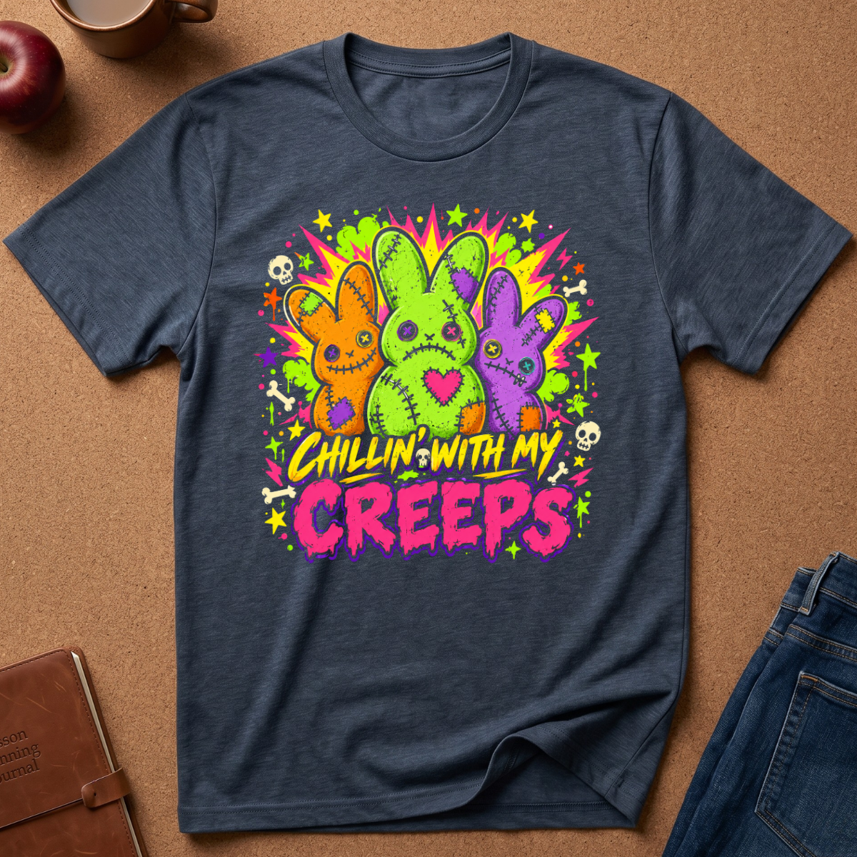 A navy blue graphic t-shirt laid flat on a brown cork background, surrounded by lifestyle props like an apple, a coffee mug, a notebook, and jeans. The t-shirt features a colorful graphic of three stitched bunny characters in orange, green, and purple, with the text 'Chillin' with my Creeps' in yellow and pink.