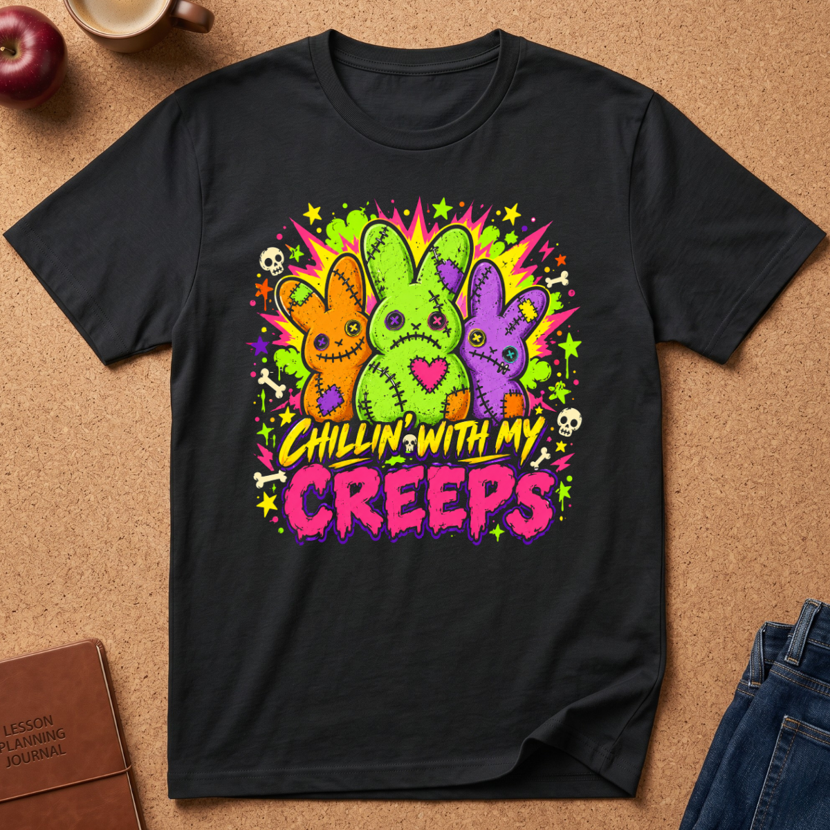 A black short-sleeve graphic T-shirt laid flat on a cork-like surface, featuring three colorful stitched bunny figures in orange, green, and purple with the text 'Chillin' with my creeps' in yellow and pink. Partial props around the shirt include an apple, a mug, a journal, and jeans.