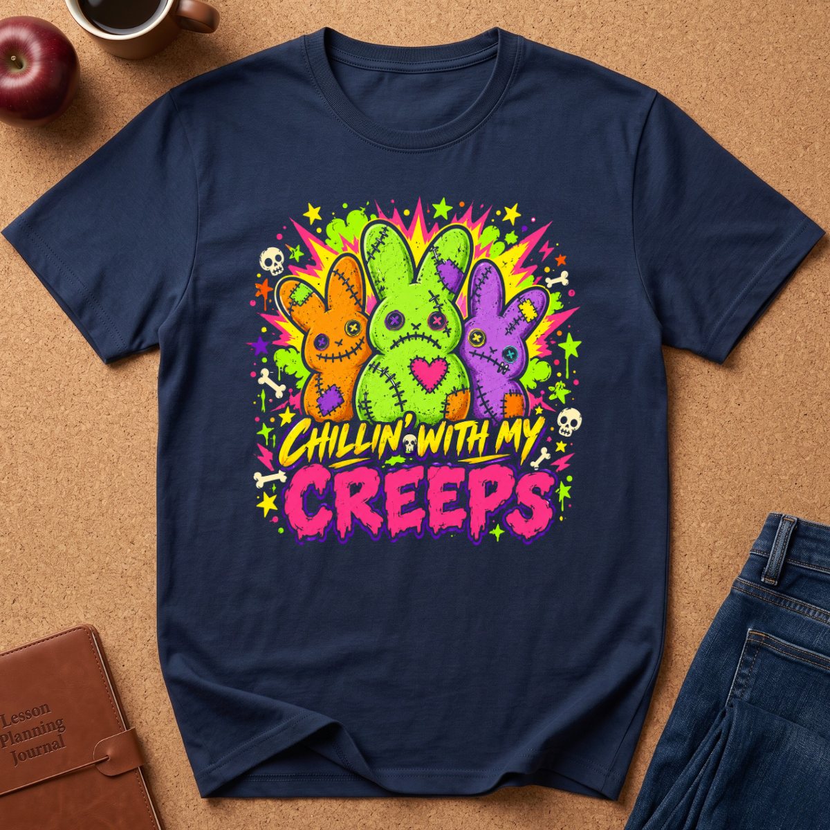 A navy blue graphic t-shirt laid flat on a brown cork background, featuring three colorful stitched bunny figures and the text 'CHILLIN' WITH MY CREEPS' in neon yellow and pink. Styled flat-lay props include an apple, a coffee cup, a journal, and jeans.