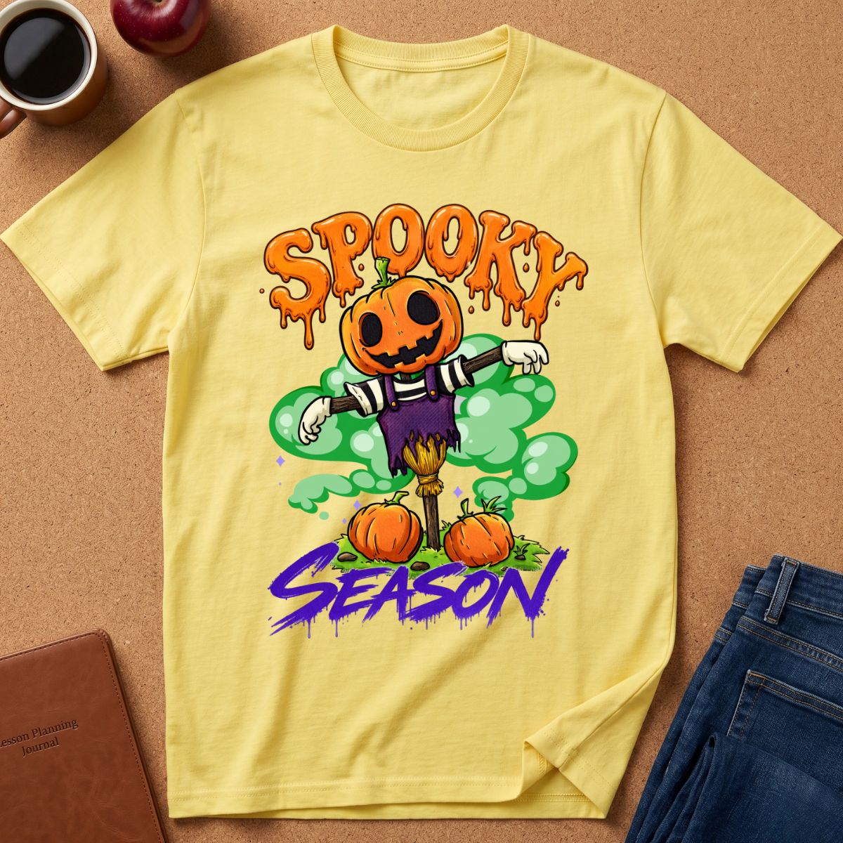 A yellow short-sleeve graphic T-shirt laid flat on a cork surface, featuring a Halloween scarecrow design with pumpkins, green smoke, and the words 'SPOOKY' and 'SEASON' in orange and purple. Surrounding props include a coffee cup, apple, notebook, and jeans.