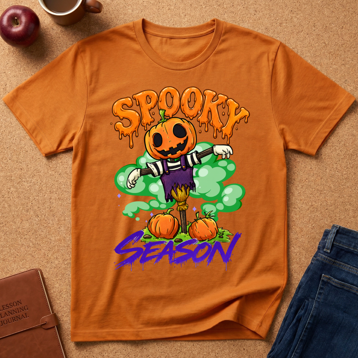 An orange crewneck t-shirt featuring a colorful Halloween graphic with a jack-o'-lantern scarecrow, pumpkins, and the text 'SPOOKY SEASON'. The shirt is displayed in a flat-lay arrangement on a cork board background alongside a brown leather journal, a red apple, a coffee mug, and blue jeans.