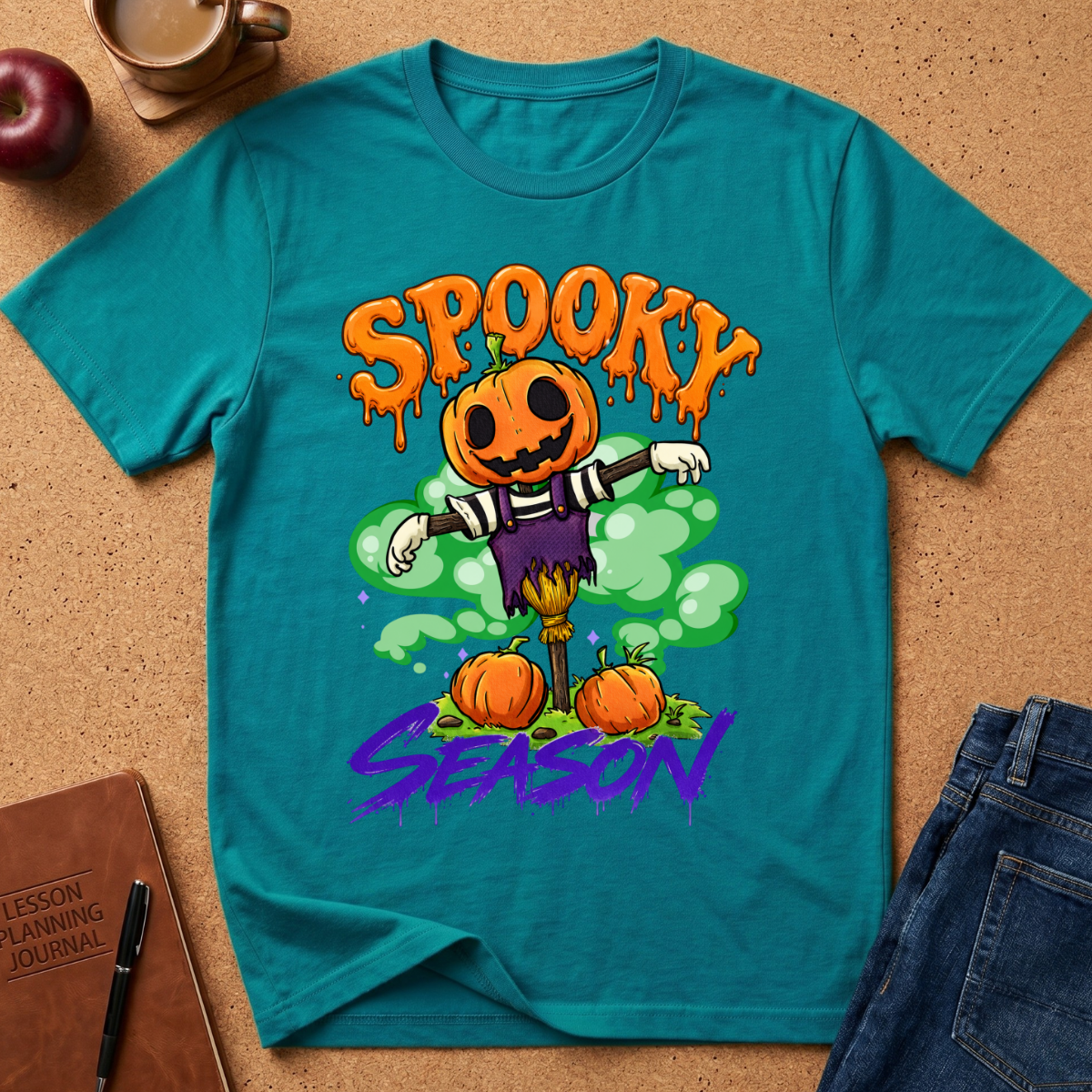 A teal short-sleeve graphic T-shirt laid flat on a cork surface, featuring a Halloween scarecrow design with pumpkins, green clouds, and the words 'Spooky Season' in orange and purple. Surrounding props include a notebook, pen, apple, coffee cup, and folded jeans.