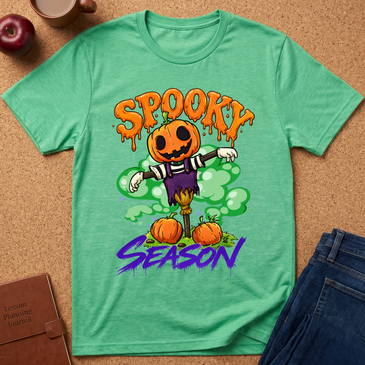 A mint green heathered graphic t-shirt laid flat on a cork background, featuring a Halloween scarecrow design with the words 'SPOOKY SEASON'. The shirt is styled with a brown leather journal, a red apple, and blue jeans.