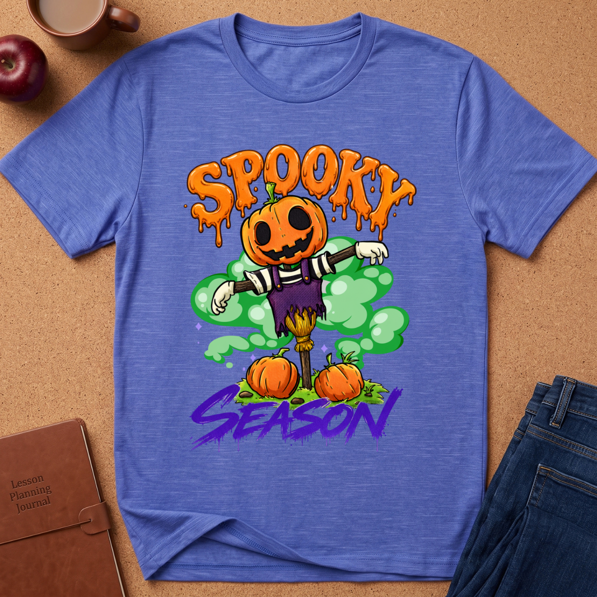 A heathered blue short-sleeve t-shirt with a colorful Halloween graphic featuring a jack-o'-lantern scarecrow, pumpkins, and the text 'SPOOKY SEASON'. The shirt is displayed in a flat-lay arrangement on a brown cork background, surrounded by a coffee mug, an apple, a notebook, and a pair of jeans.