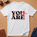 A white short-sleeve crewneck t-shirt with a graphic print reading 'YOU ARE' in black, overlaid with a red heartbeat line and red hearts, laid flat on a cork background with a coffee cup, apple, and jeans nearby.