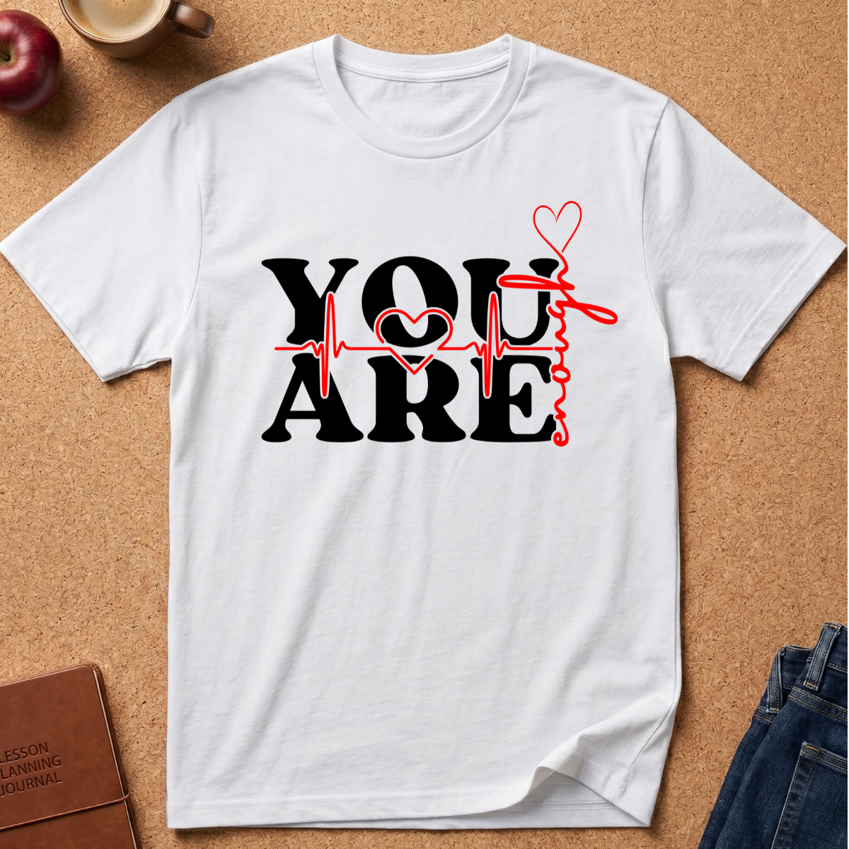 A white short-sleeve crewneck t-shirt with a graphic print reading 'YOU ARE' in black, overlaid with a red heartbeat line and red hearts, laid flat on a cork background with a coffee cup, apple, and jeans nearby.