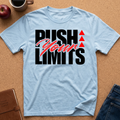 A light blue crewneck t-shirt laid flat on a cork background, featuring a graphic print that reads 'PUSH YOUR LIMITS' in black and red script, with three red triangles. A coffee mug, apple, notebook, and jeans are styled around it.