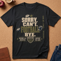 A black graphic t-shirt laid flat on a cork board background, featuring a football-themed design with the text 'SORRY, CAN'T FOOTBALL, BYE.' in distressed beige and olive green lettering, accompanied by footballs, a crown, and a shield crest.