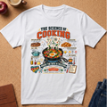 A white graphic t-shirt laid flat on a cork background, featuring a colorful infographic design titled 'THE SCIENCE OF COOKING' with illustrations of food, cookware, and cooking terms. Styled flat-lay props include an apple, a coffee cup, a notebook, and jeans.