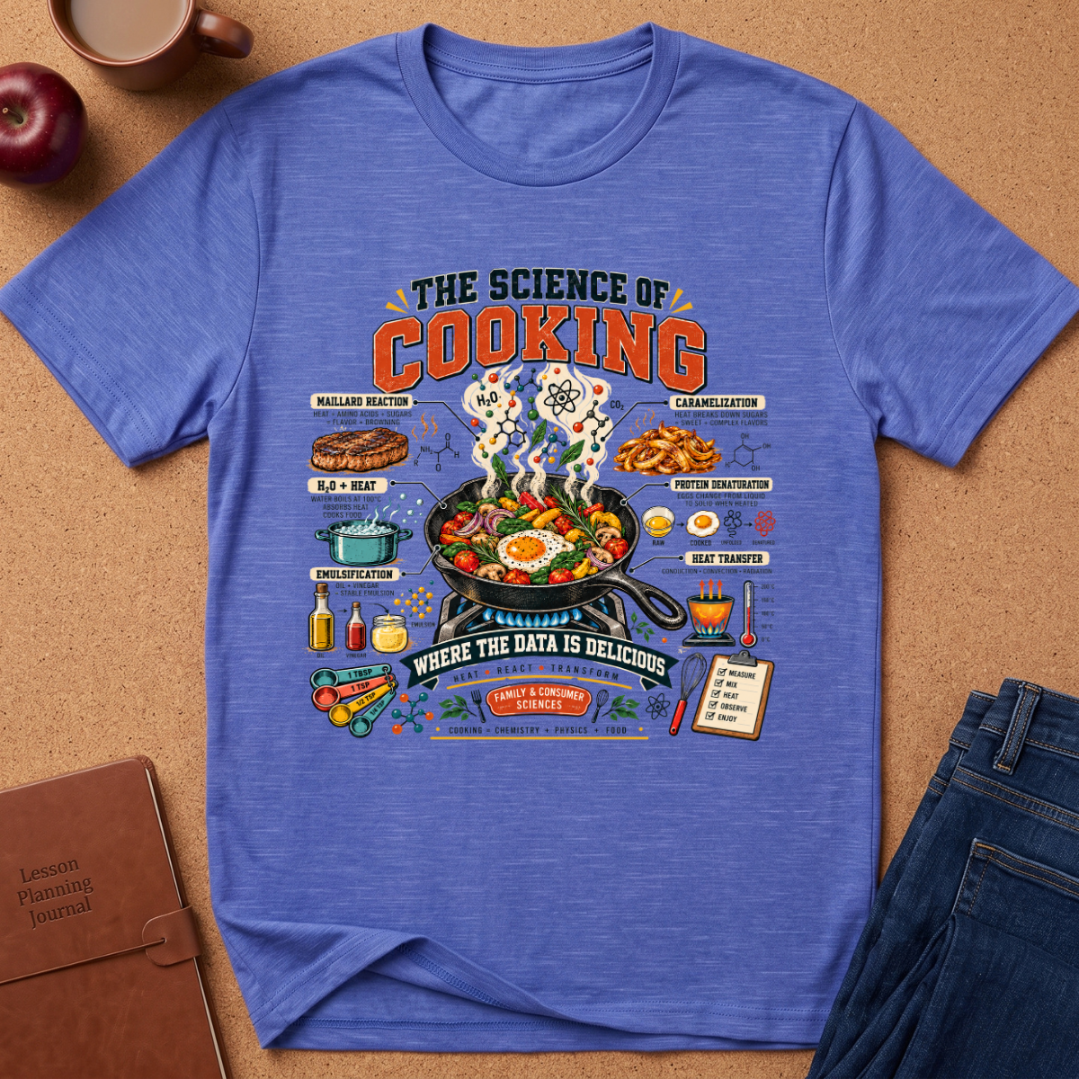 A heathered blue graphic t-shirt laid flat on a brown cork background, surrounded by lifestyle props including an apple, a coffee cup, a notebook, and a pair of jeans. The t-shirt features a detailed cooking-themed graphic with illustrations of food, ingredients, and text like 'THE SCIENCE OF COOKING' and 'WHERE THE DATA IS DELICIOUS'.