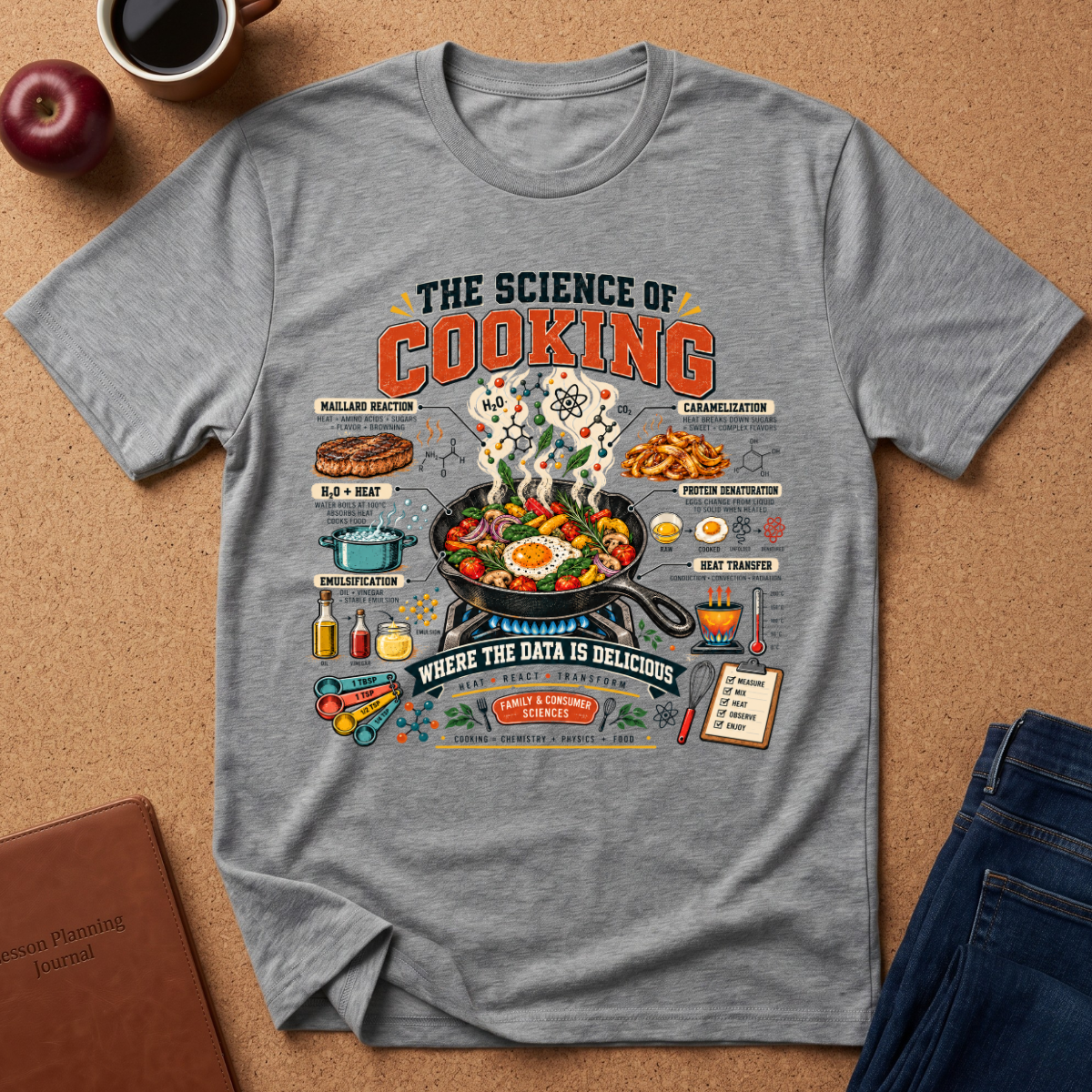 A heather gray graphic T-shirt laid flat on a cork surface, surrounded by lifestyle props including an apple, a coffee cup, a notebook, and jeans. The shirt features a colorful retro-style cooking infographic with the text 'THE SCIENCE OF COOKING' and illustrated food preparation elements.