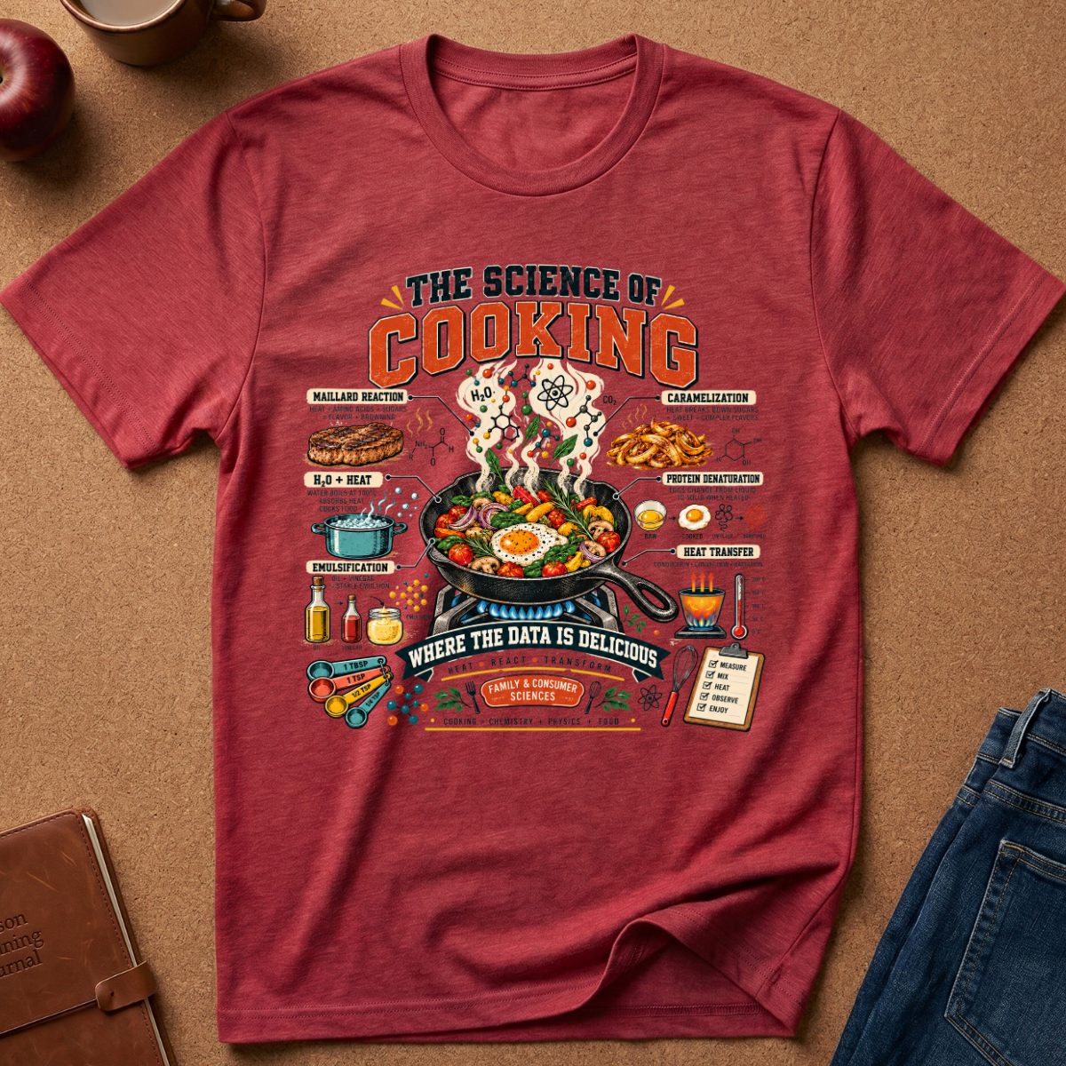 A heathered red graphic t-shirt laid flat on a brown cork background, surrounded by lifestyle props including an apple, a coffee cup, a notebook, and a pair of jeans. The t-shirt features a detailed cooking-themed graphic with illustrations of food, ingredients, and text like 'THE SCIENCE OF COOKING' and 'WHERE THE DATA IS DELICIOUS'.