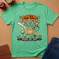 A mint green graphic t-shirt laid flat on a cork background, styled with an apple, coffee cup, brown journal, and blue jeans. The t-shirt features a colorful retro-style graphic that reads 'BAKING IS CHEMISTRY YOU CAN EAT' with illustrations of baking ingredients and utensils.