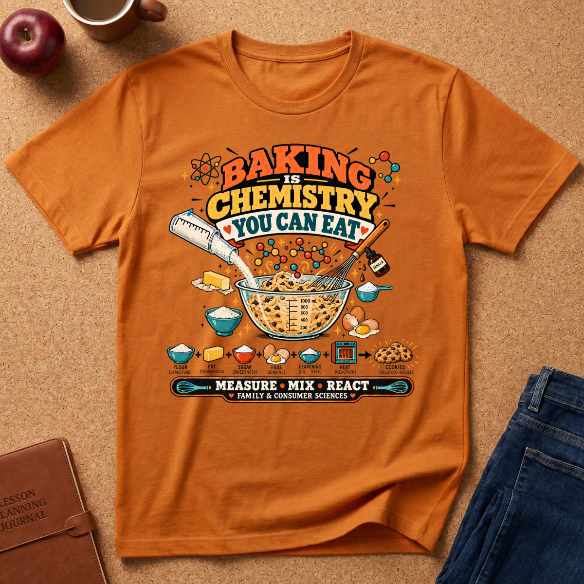 Orange graphic t-shirt laid flat on a cork background, styled with an apple, coffee cup, brown journal, and blue jeans. The t-shirt features a colorful retro-style graphic that reads 'BAKING IS CHEMISTRY YOU CAN EAT' with illustrations of baking ingredients and utensils.
