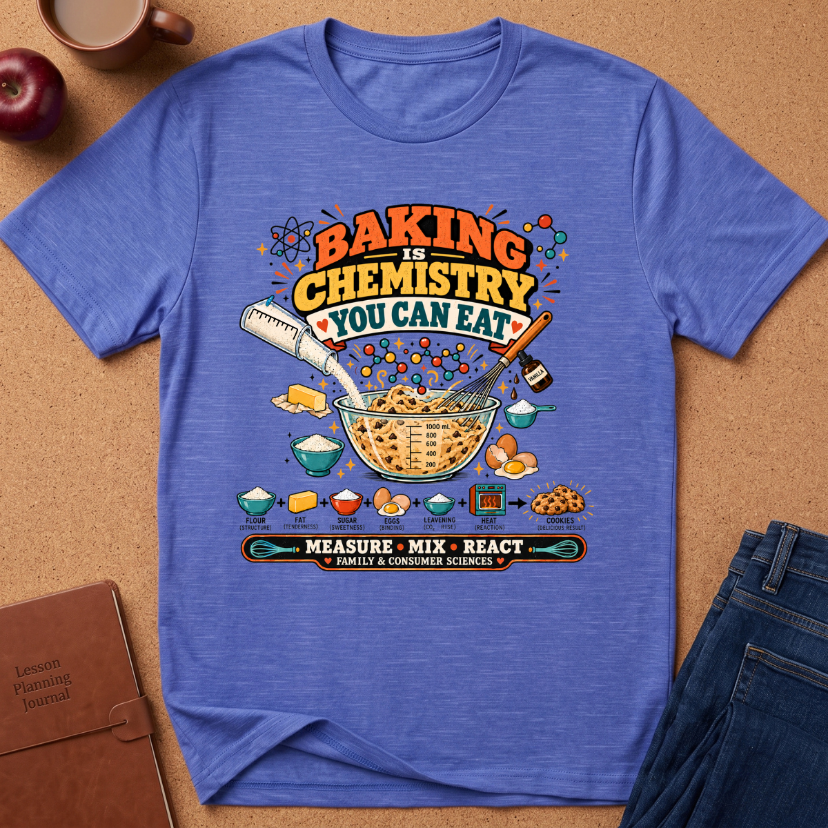 A heather blue graphic t-shirt laid flat on a brown cork background, surrounded by lifestyle props including an apple, a coffee cup, a notebook, and a pair of jeans. The t-shirt features a colorful 'Baking is Chemistry You Can Eat' graphic with baking ingredients and the words 'Measure Mix React'.
