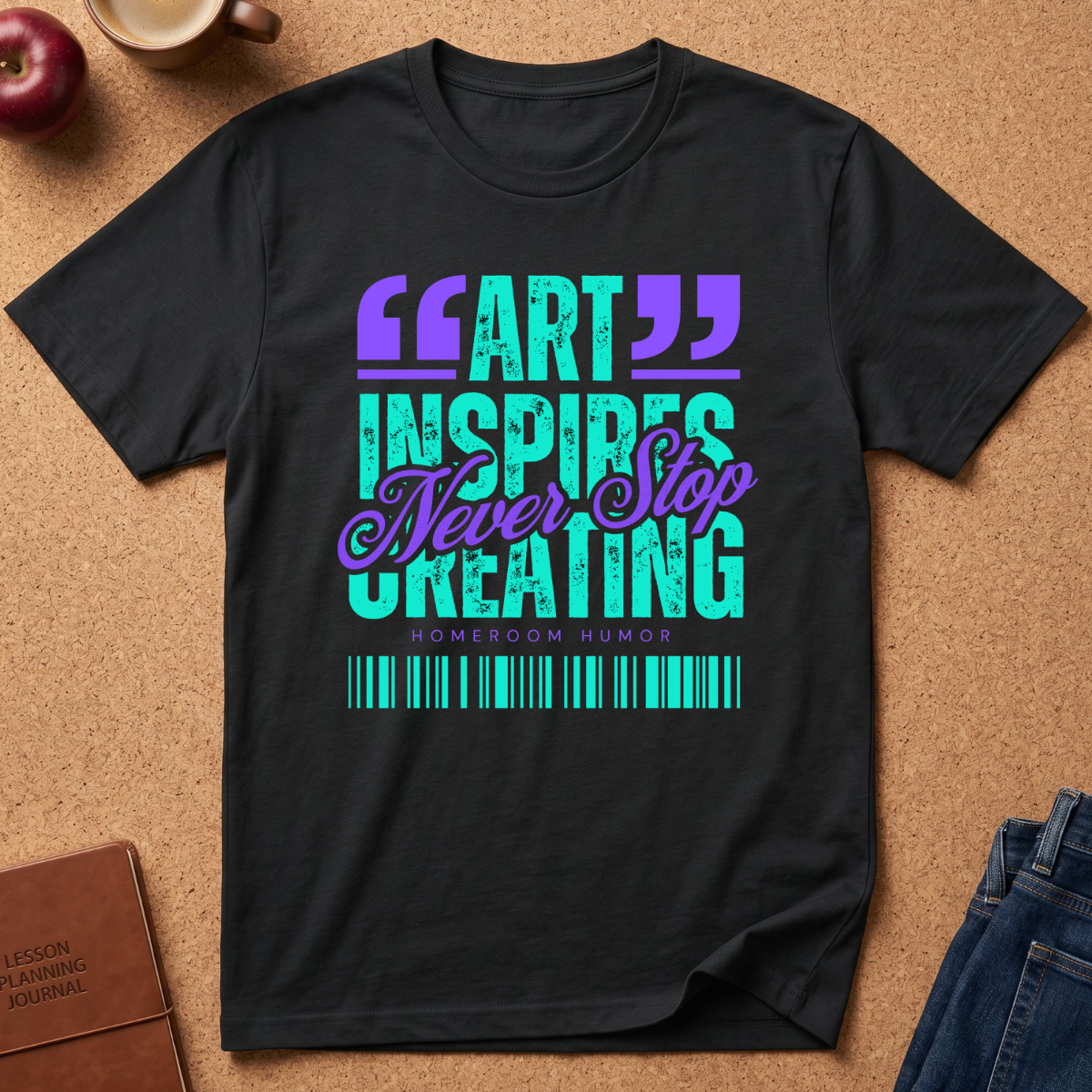 A black short-sleeve graphic T-shirt laid flat on a cork surface, featuring the phrase 'ART INSPIRES Never Stop CREATING' in purple and teal text, with 'HOMEROOM HUMOR' and a barcode graphic below. Surrounding props include an apple, a coffee cup, a brown journal, and blue jeans.