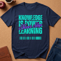 A navy blue graphic t-shirt laid flat on a brown cork board background, surrounded by lifestyle props including an apple, a coffee cup, a leather journal, and blue jeans. The t-shirt features a colorful text graphic that reads 'KNOWLEDGE IS POWER Never Stop LEARNING' with a barcode design at the bottom.