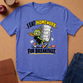 A heathered blue graphic t-shirt laid flat on a cork background, featuring a cartoon green creature carrying a stack of papers and the text 'I EAT HOMEWORK FOR BREAKFAST'. Surrounding props include an apple, a coffee mug, a notebook, and jeans.
