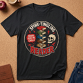 A black graphic t-shirt laid flat on a cork background, featuring a circular design with the words 'SPINE-TINGLING READER' and 'BOOKS GIVE ME CHILLS' around a stack of books, a skull, and red lightning accents. Styled flat-lay props include an apple, a coffee cup, a journal, and jeans.