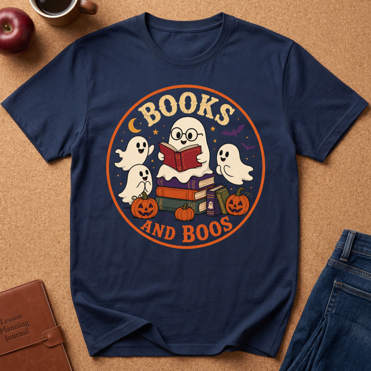 A navy blue graphic t-shirt laid flat on a cork background, featuring a circular Halloween-themed design with the words 'BOOKS AND BOOS' and illustrations of ghosts, books, pumpkins, and stars. Styled flat-lay props include an apple, a coffee cup, a brown journal, and blue jeans.