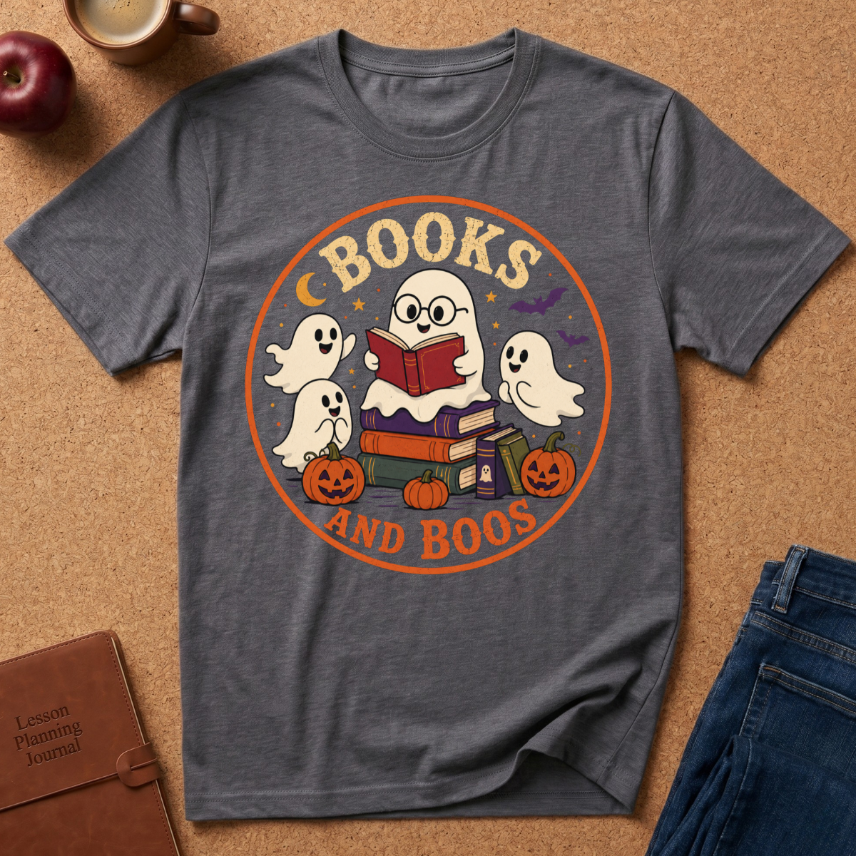 A dark gray graphic t-shirt laid flat on a cork background, featuring a circular Halloween design with ghosts, books, pumpkins, and the words 'BOOKS AND BOOS'. Styled flat-lay props include an apple, a coffee cup, a brown journal, and blue jeans.