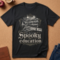 A black short-sleeve graphic T-shirt laid flat on a cork surface, featuring a white illustrated design of stacked books, a skull, bones, and the text 'The Art of Spooky education.' Surrounding props include an apple, a cup, a journal, and folded blue jeans.