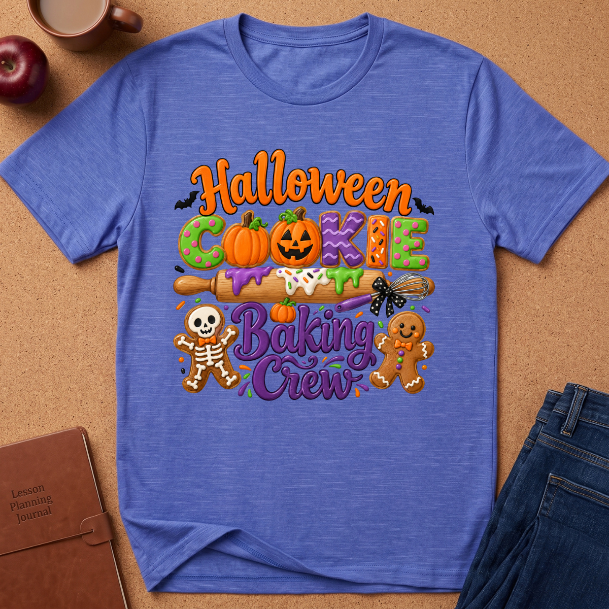 A heathered blue crewneck t-shirt with a colorful 'Halloween Cookie Baking Crew' graphic, styled in a flat-lay on a cork background with a notebook, apple, coffee cup, and jeans.