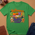 A green short-sleeve crewneck t-shirt with a colorful 'Halloween Cookie Baking Crew' graphic, laid flat on a brown cork background with lifestyle props including a coffee mug, an apple, a journal, and jeans.