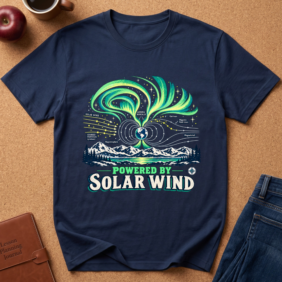 A navy blue graphic t-shirt laid flat on a brown cork board background, styled with an apple, a coffee cup, a journal, and jeans. The t-shirt features a colorful graphic of the aurora borealis over mountains and solar wind diagrams with the text 'POWERED BY SOLAR WIND'.
