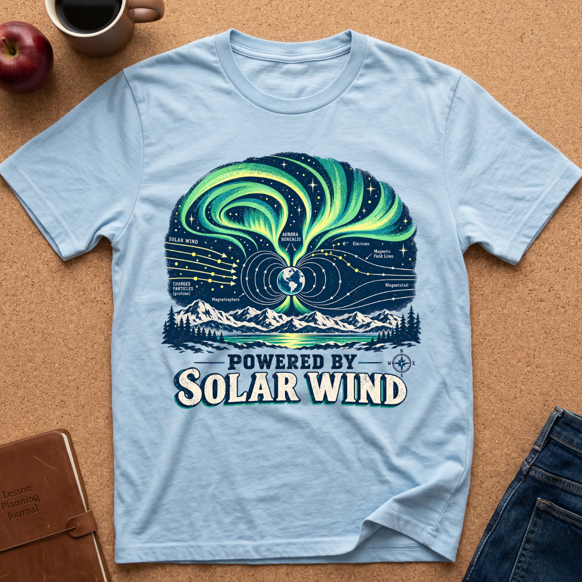 A light blue graphic T-shirt laid flat on a cork surface, surrounded by a coffee mug, apple, notebook, and jeans. The shirt features a large illustrated design with aurora-like green and blue swirls, stars, mountains, and the text 'POWERED BY SOLAR WIND'.
