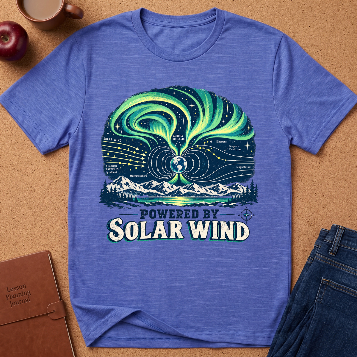 A heathered blue short-sleeve t-shirt laid flat on a cork surface, featuring a large graphic print of the aurora borealis over mountains and stars with the text 'POWERED BY SOLAR WIND.' A brown notebook, apple, coffee cup, and folded jeans are placed around it as styling props.