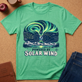A green short-sleeve graphic T-shirt laid flat on a cork surface, with an illustrated aurora and mountain scene and the text 'POWERED BY SOLAR WIND.' Jeans, an apple, a coffee cup, and a notebook are partially visible around it as styling props.