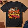 A black short-sleeve graphic T-shirt laid flat on a cork background, featuring autumn-themed text reading 'Thankful for Thanksgiving Break' with leaves, pumpkins, an apple, a mug, and a chalkboard sign. No person or real environment is present in the sense of a lifestyle scene; this is a styled flat-lay product shot.