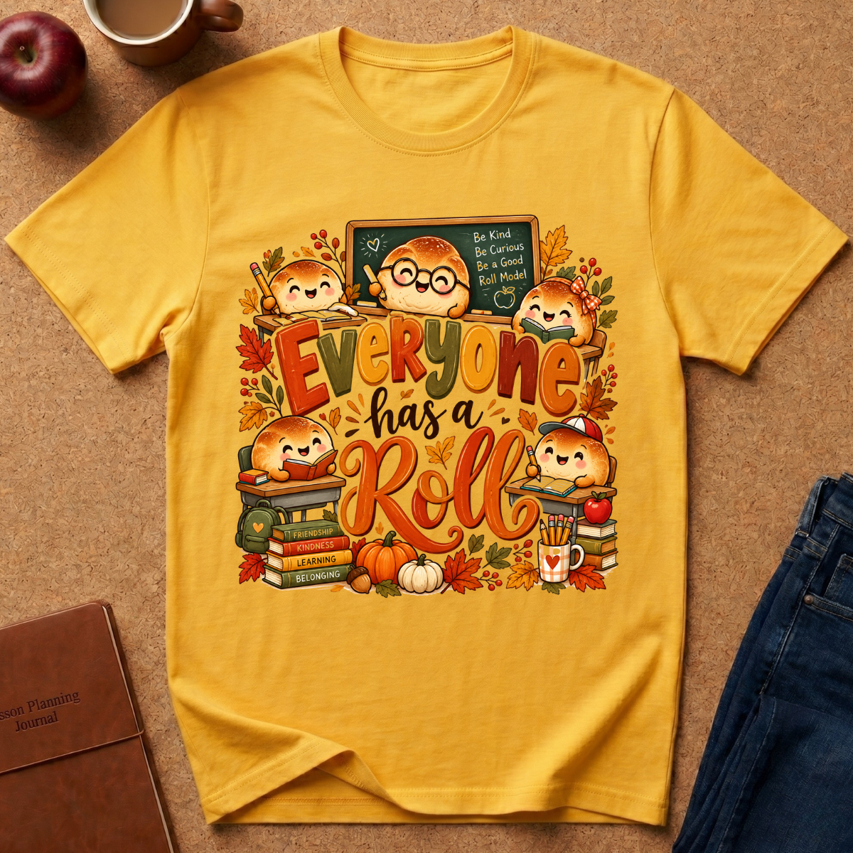 A yellow short-sleeve graphic T-shirt laid flat on a cork-like surface, surrounded by autumn-themed props including an apple, mug, notebook, and jeans. The shirt features a colorful cartoon school-bunny illustration with the phrase 'Everyone has a Roll' in large fall-colored letters.