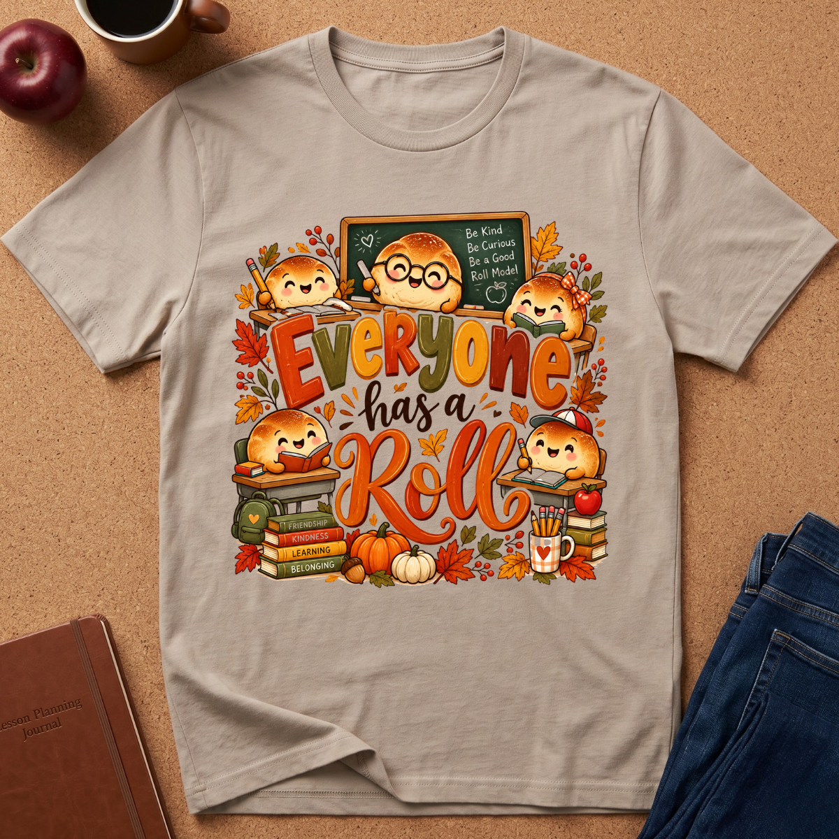 A light beige graphic t-shirt laid flat on a brown cork background, surrounded by autumn-themed props like an apple, a coffee mug, a notebook, and blue jeans. The t-shirt features a colorful cartoon illustration of cute bread characters in glasses, books, pumpkins, and fall leaves, with the text 'Everyone has a Roll'.