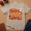 A light beige graphic t-shirt laid flat on a brown cork background, featuring a colorful autumn-themed design with the text 'Running on Pumpkin Spice' surrounded by pumpkins, leaves, and a coffee cup illustration. Styled flat-lay props include an apple, a notebook, a coffee mug, and blue jeans.