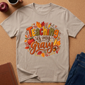 A light beige graphic t-shirt laid flat on a cork background, featuring the phrase 'Teaching is my Gray' in a colorful autumn-themed design with leaves, an apple, and a pencil. Styled flat-lay props include an apple, a coffee cup, a notebook, and blue jeans.