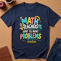 A navy blue graphic t-shirt laid flat on a brown cork background, surrounded by lifestyle props including an apple, a coffee cup, a leather journal, and blue jeans. The t-shirt features a colorful graphic that reads 'IT MATH TEACHERS HAVE TOO MANY PROBLEMS' with school-themed illustrations.